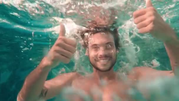 Μέτριο πλάνο του νεαρού ελκυστικό άνδρα με μεγάλο χαμόγελο δείχνει αντίχειρες-up και καταδύσεις κάτω από το νερό - Πλάνα, βίντεο