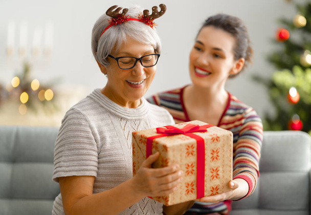 Merry Christmas and Happy Holidays! Senior mom and her adult daughter exchanging gifts. Having fun near tree indoors. Loving family with presents in room.  - Foto, Imagem