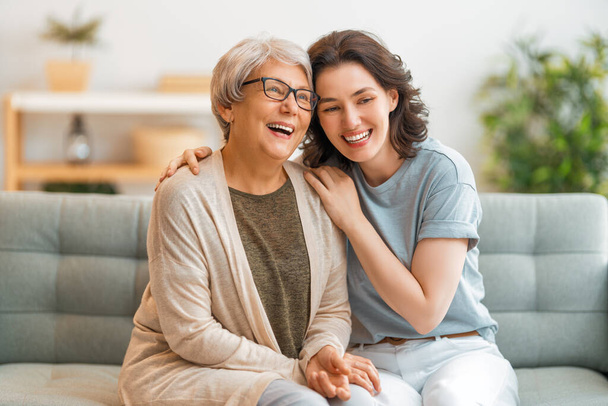 Beautiful mother and daughter are talking and smiling while sitting on couch at home. - 写真・画像
