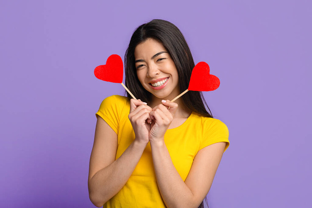 Playful asian woman posing with two paper hearts in hands, purple background - Photo, Image