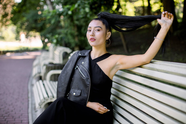 Outdoor summer lifestyle portrait of romantic young beautiful Asian woman in casual stylish black outfit, sitting on a bench in green public park, touching her ponytail, looking at camera - Valokuva, kuva
