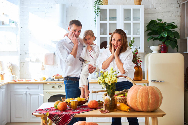 A happy family of three, mother, father and daughter, stand on a light kitchen, a wooden table with an autumn harvest - pumpkins, apples - Fotoğraf, Görsel