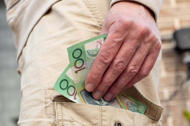 Hand hält australische 100-Dollar-Banknoten in der Tasche der Cargo-Shorts. Konzept zur Handelsfinanzierung. - Foto, Bild