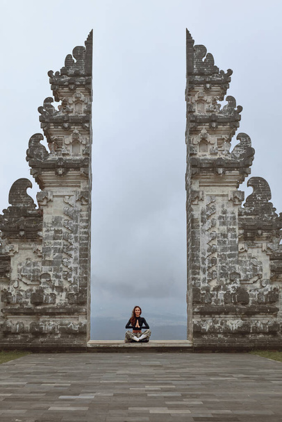 Beautiful Girl In Lotus Pose Meditating Near Gates Of Heaven In Pura Lempuyang Temple In Bali, Indonesia. Calm And Peaceful Young Woman Sitting With Crossed Legs Near Ancient Architecture, Asia. - Photo, Image