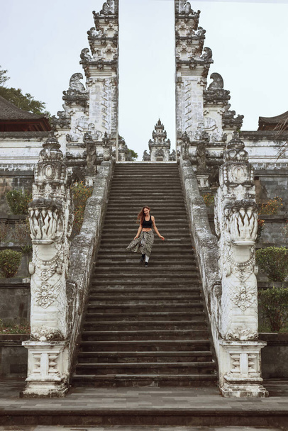 Beautiful Girl Going Down Stone Stairs From Gates Of Heaven In Pura Lempuyang Temple In Bali, Indonesia. Happy Young Woman Explores Famous Landmark In Asia. - Photo, Image