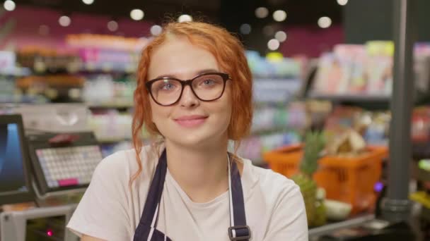 Ritratto di graziosa cassiera in occhiali che lavora dietro la cassa cercando felicemente nella macchina fotografica nel moderno negozio di alimentari - Filmati, video