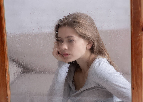 Hermosa joven sintiéndose triste por el clima lluvioso, mirando por la ventana y pensando - Foto, Imagen