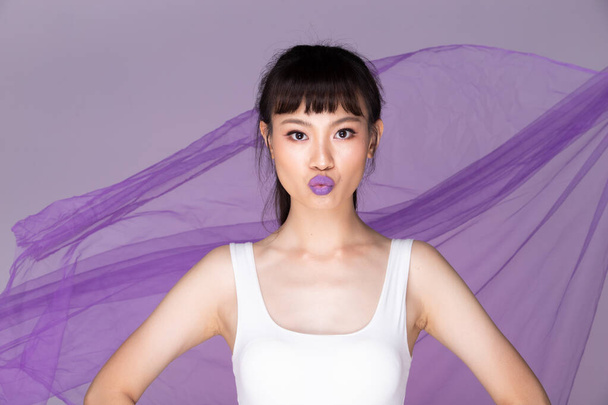 Portrait of Fashion 20s Asian Woman has beautiful Make Up white vast, strong feeling poses over purple Background and throwing fluttering clothes fabric in air - Foto, Imagem