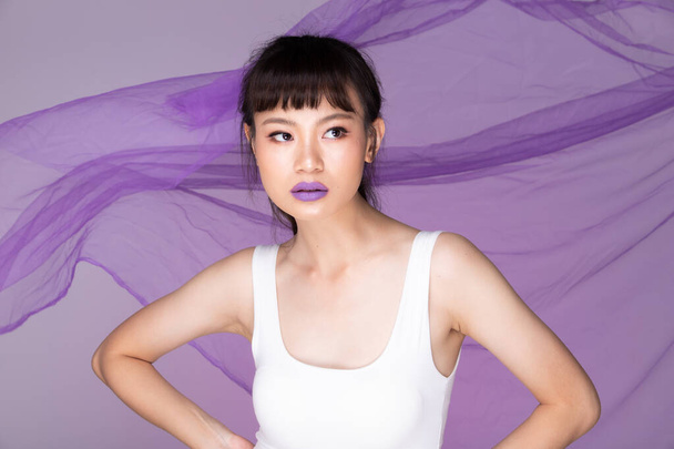 Portré divat 20-as ázsiai nő gyönyörű Make Up fehér hatalmas, erős érzés pózol a lila háttér és dobott lebegő ruha szövet a levegőben - Fotó, kép