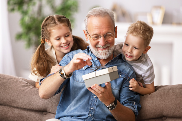幸せな家族高齢者祖父と孫の女の子と男の子オープニングギフトボックスながら休日のお祝いの間にソファに休みます-ホーム - 写真・画像