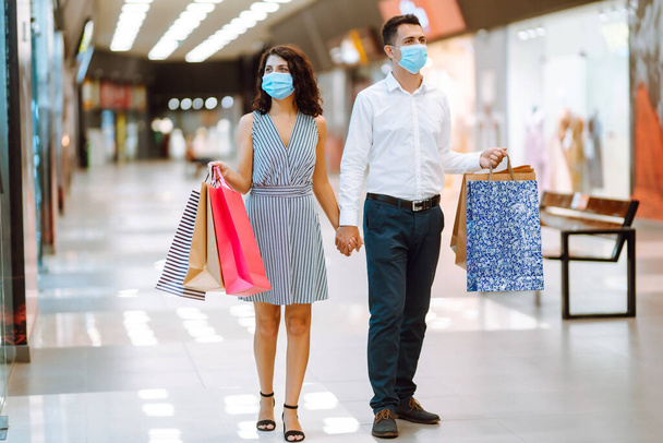 Young loving couple in protective medical mask with shopping bags walking in the mall. Shopping during the coronavirus epidemic. Sale, consumerism, Black friday concept.  - Фото, изображение
