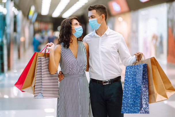 Young loving couple in protective medical mask with shopping bags walking in the mall. Shopping during the coronavirus epidemic. Sale, consumerism, Black friday concept.  - Foto, imagen
