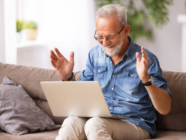 Expressive bearded elderly male relaxing on sofa and browsing laptop on weekend day at hom - 写真・画像