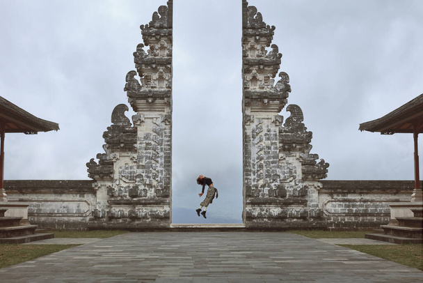 Beautiful Girl Full-Length Portrait Near Gates Of Heaven In Pura Lempuyang Temple In Bali, Indonesia. Happy Young Woman Doing Back Band Jump Near Ancient Architecture In Asia. - Photo, Image