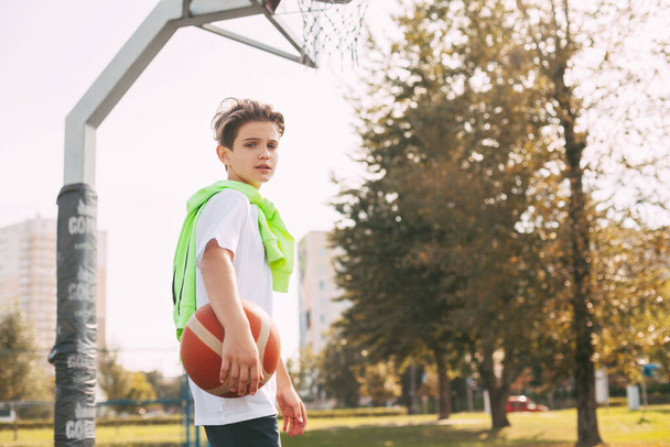 Красивый подросток держит в руках баскетбольный мяч и смотрит в камеру. Портрет мальчика с мячом в руках. Спорт, образование, досуг - Фото, изображение