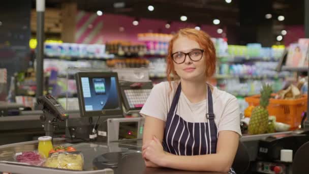 Fiatal, csinos, mosolygós pénztáros szemüvegben és csíkos kötényben boldogan pózol a kamera előtt, miközben a pénztárgép mögött dolgozik a modern élelmiszerboltban. - Felvétel, videó