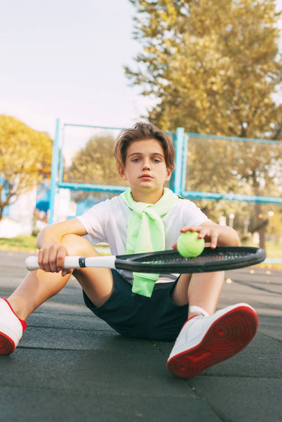 Χαριτωμένος έφηβος κάθεται στο γήπεδο του τένις και ξεκουράζεται μετά από μια προπόνηση. Αθλητισμός, αθλητισμός, τρόπος ζωής, αναψυχή - Φωτογραφία, εικόνα