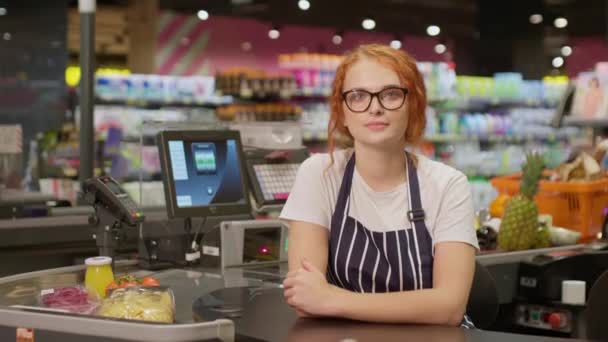 Muotokuva nuori hymyilevä kassanhoitaja silmälasit ja raidallinen esiliina tarkkaavaisesti kameran ja hymyillen työskentelee moderni ruokakauppa - Materiaali, video