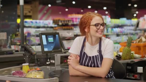 Elég vidám vörös pénztáros szemüvegben és csíkos kötényben boldogan pózol a kamera előtt, miközben a modern szupermarketben pénztárgép mögött dolgozik. Az élelmiszerbolt koncepciója - Felvétel, videó