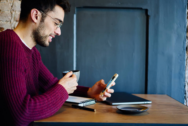 Вид сбоку веселый молодой бородатый мужчина в повседневной одежде сидит за столом возле стены и просматривает смартфон во время питья кофе - Фото, изображение