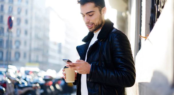 Positive bearded male in stylish outfit standing on street near modern building with cup of coffee and browsing mobile phone during walk - Foto, Imagem