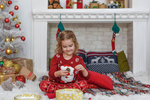 Merry Christmas and Happy Holidays! Cute little child girl is decorating the Christmas tree indoors. Happy little smiling girl with christmas gift box. - Foto, Bild