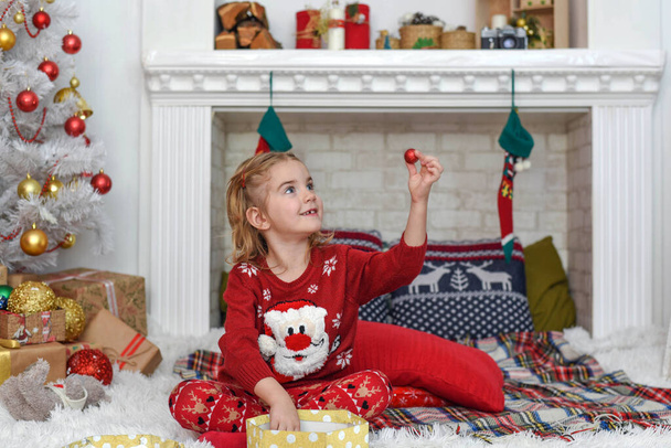 Merry Christmas and Happy Holidays! Cute little child girl is decorating the Christmas tree indoors. Happy little smiling girl with christmas gift box. - Foto, afbeelding
