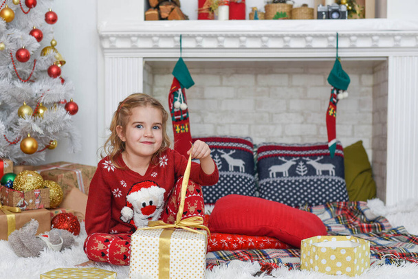 Boldog Karácsonyt és Kellemes Ünnepeket! Aranyos kislány díszíti a karácsonyfát odabent. Boldog kis mosolygós lány karácsonyi ajándékdobozzal. - Fotó, kép
