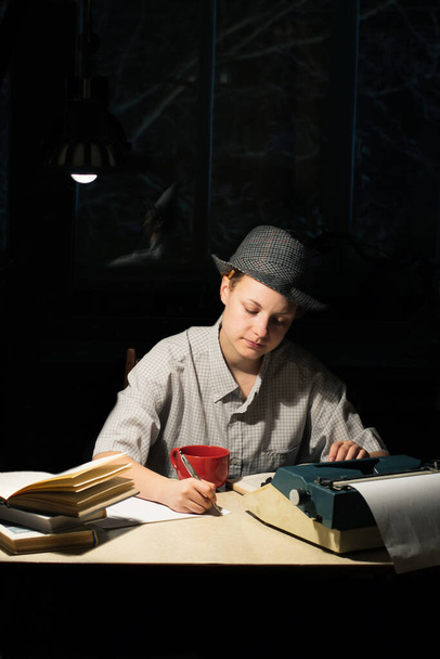 Porträt eines Mädchens mit Hut, das mit Schreibmaschine und Büchern an einem Tisch sitzt und nachts Notizen macht - Foto, Bild