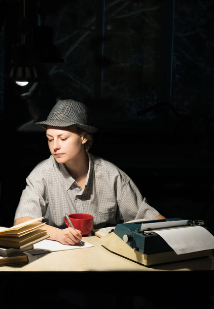Portret van een meisje in een hoed zittend aan een tafel met een schrijfmachine en boeken, 's nachts notities makend - Foto, afbeelding