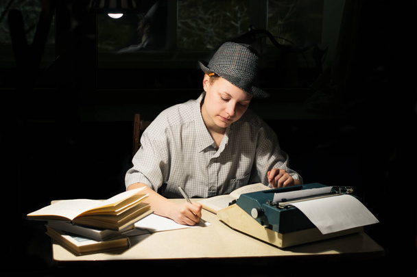 Portrait d'une fille dans un chapeau assis à une table avec une machine à écrire et des livres, prenant des notes la nuit - Photo, image