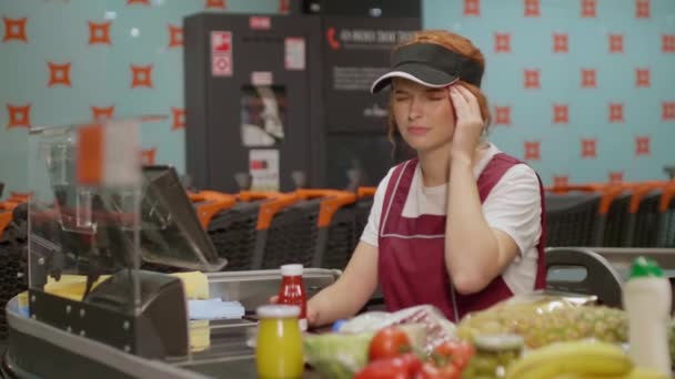Junge seriöse Kassiererin in Uniform massiert Schläfen, während sie im modernen Supermarkt arbeitet. Verkäuferin arbeitet hinter Kasse - Filmmaterial, Video