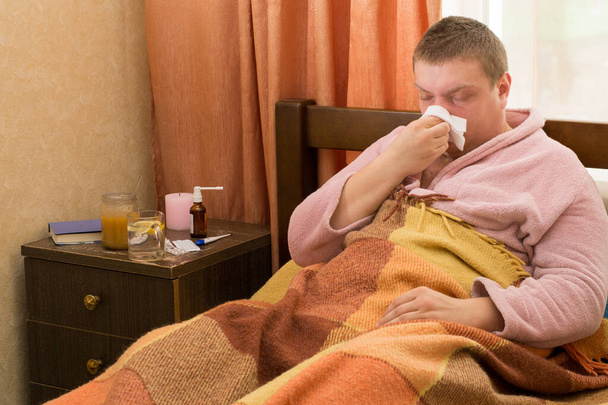 Sick guy in a bathrobe, lying in the bed and blowing - Photo, Image