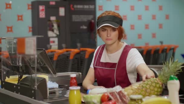 Junge, seriöse Kassiererin in Uniform, die in einem modernen Supermarkt arbeitet. Unzufriedene Verkäuferin bei der Arbeit im Lebensmittelgeschäft - Filmmaterial, Video