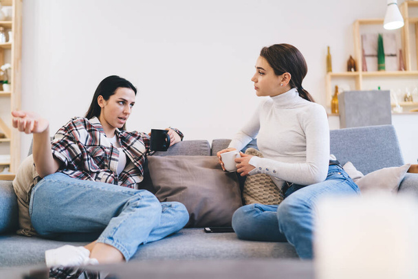 Female friends in casual wearing chatting with each other while sitting on sofa and drinking coffee in cozy living room at home - Foto, imagen