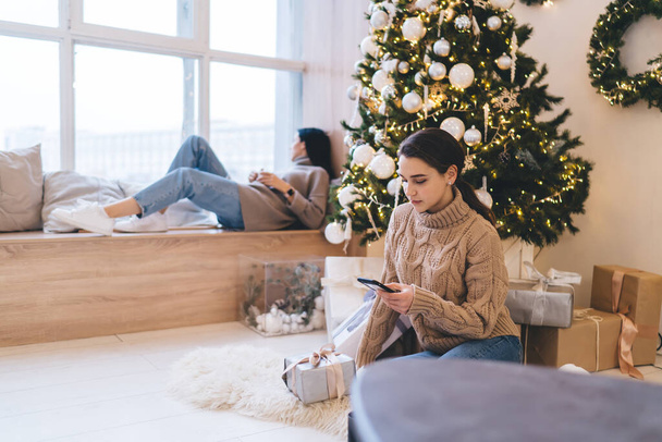 Serious woman browsing smartphone while sitting on floor near Christmas tree with female friend lying near window during winter vacation - Foto, imagen