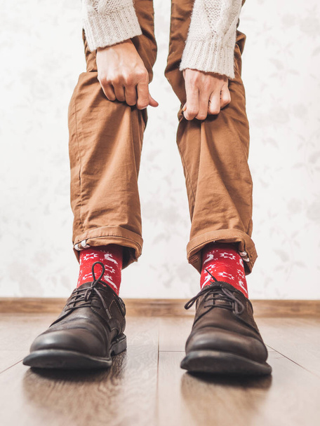 Young man pulls up leg of his chinos trousers to show bright red socks with reindeers on them. Scandinavian pattern. Winter holiday spirit. Casual outfit for New Year and Christmas celebration. - 写真・画像