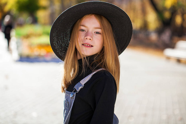 Portrait of a red-haired little girl in a black hat walks in the autumn park - Fotoğraf, Görsel