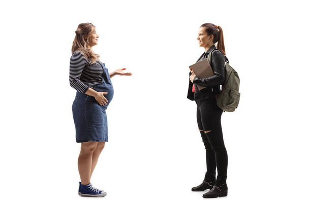 Full length profile shot of a young pregnant woman talking to a female student with a backpack and books isolated on white background - Fotó, kép