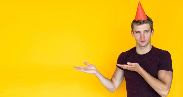 young man on a yellow background. birthday concept with a cap on the head, points with two palms to an empty space for text - Φωτογραφία, εικόνα
