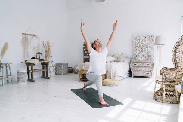 Cheerful caucasian male in perfect body shape keeping fit during morning training on carpet and cozy home interior, smiling hipster guy satisfied with practicing and training standing in asana - Valokuva, kuva