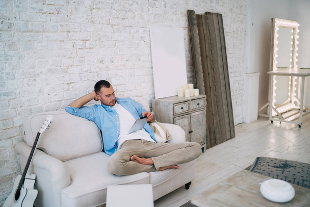 Full body man wearing casual clothes and eyeglasses sitting on comfortable couch and browsing tablet during break in cozy living room - Foto, imagen