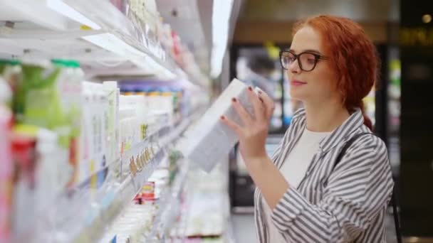Bella ragazza rossa sorridente in occhiali vestiti casual usura scegliendo yogurt nel moderno negozio di alimentari. Concetto supermercato - Filmati, video