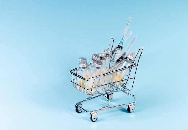 Medical syringes and glass vials of vaccine in a metal shopping cart, on a blue background. The concept of vaccination of the population. Close-up, selective focus - Foto, Bild