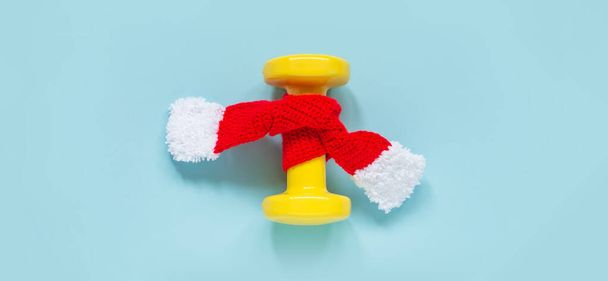 Exercise, fitness and development of the concept of a happy Christmas and a happy New year. Yellow dumbbell in a red knitted scarf on a blue background. Banner. Top view, copy space - Φωτογραφία, εικόνα