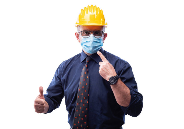 Engineer isolated on white background wears surgical mask to prevent Coronavirus spread, makes OK sign with thumb up. Preventing Pandemic Covid-19 at the workplace. - 写真・画像