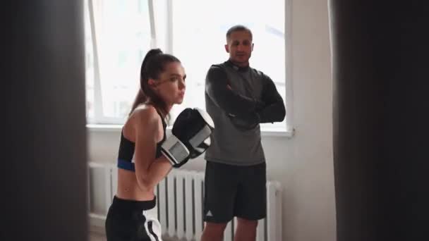 Een strenge trainer kijkt toe hoe zijn vrouwelijke kickbokser student een stomp op een bokszak oefent in een ruime trainingsruimte - Video