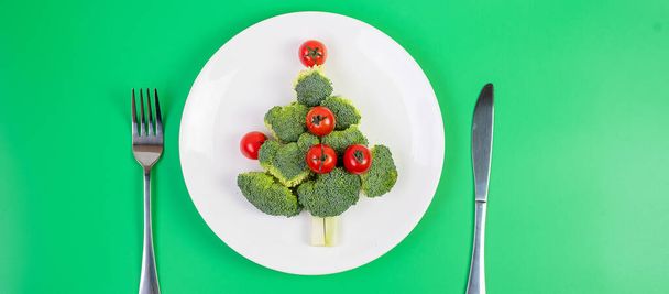 Beyaz tabakta organik sebzelerden oluşan Noel ağacı; Brokoli ve yeşil arka planda çatal ve bıçaklı domatesler. Mutlu Noeller ve mutlu yeni yıl partisi, yeni sen, sağlıklı yemek ve vejetaryen konsepti - Fotoğraf, Görsel