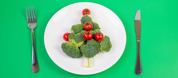白いプレート上の有機野菜のクリスマスツリー;緑の背景にフォークとナイフでブロッコリーとトマト。メリークリスマスと幸せな新年のパーティー,新しいあなた,健康食品やベジタリアンの概念 - 写真・画像