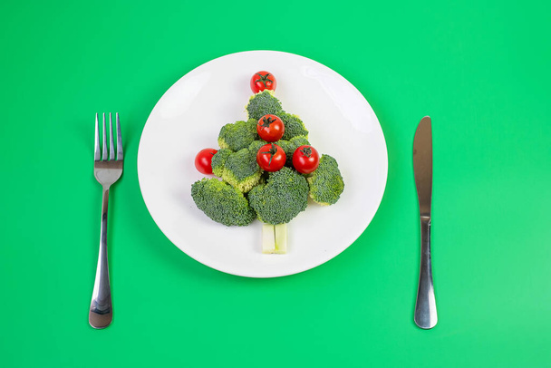 Choinka z ekologicznych warzyw na białym talerzu; Brokuły i pomidory z widelcem i nożem na zielonym tle. Wesołych Świąt i szczęśliwego Nowego Roku, New You, Zdrowe jedzenie i koncepcja wegetariańska - Zdjęcie, obraz